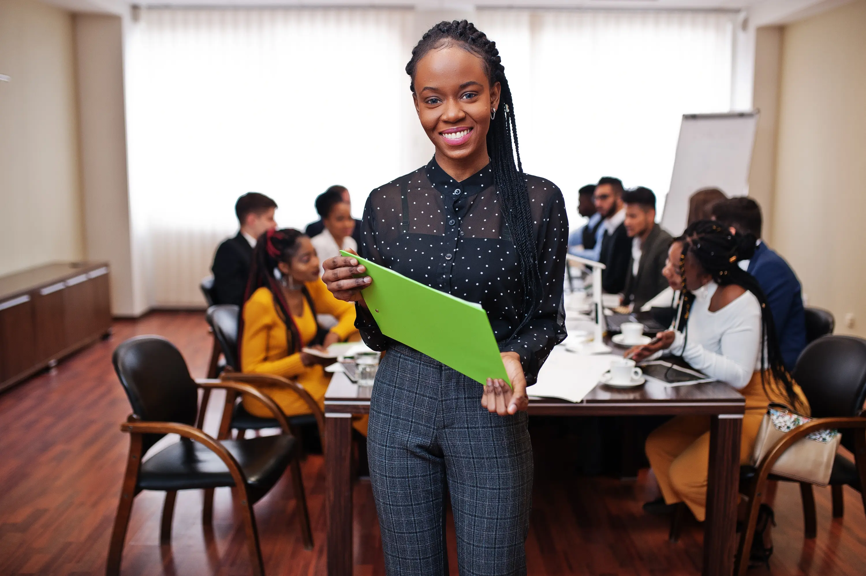 A smiling young professional holding a folder, with her colleagues in a meeting behind her